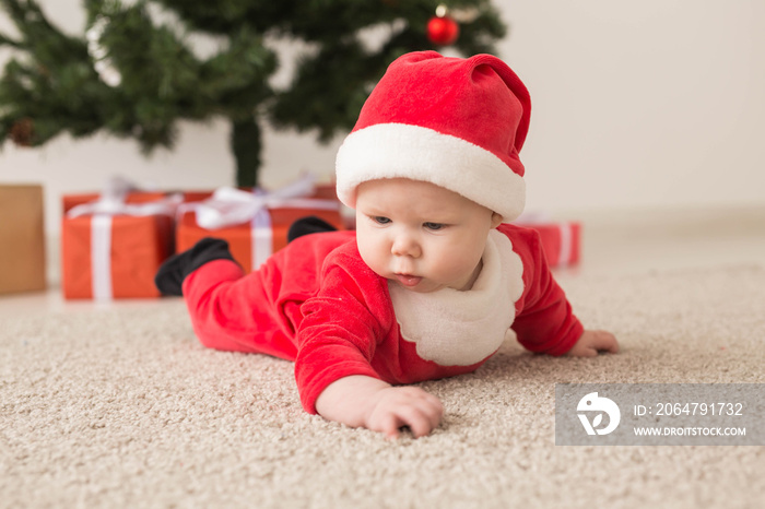 Cute baby girl wearing santa claus suit crawling on floor over Christmas tree. Holiday season.