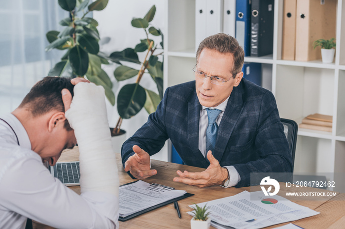 worker with broken arm sitting at table and reading documents opposite to businessman in blue jacket