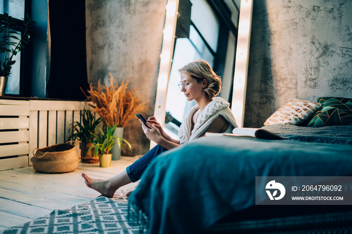 Woman in glasses sitting near bed and using smartphone