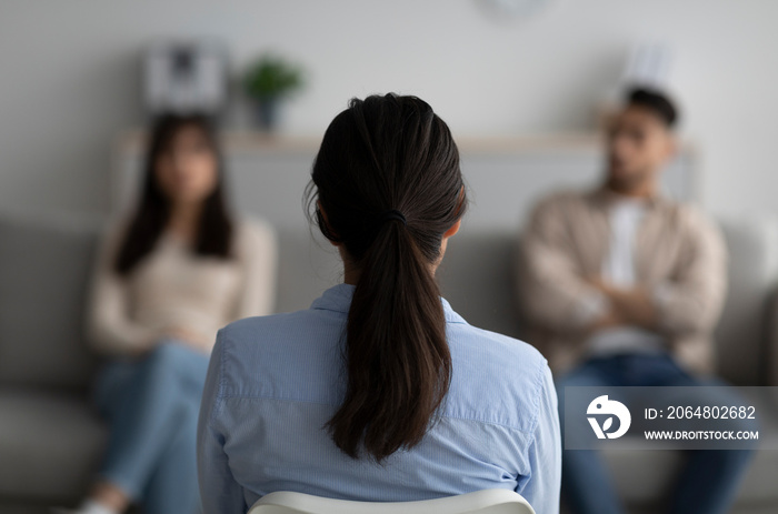 Female psychologist sitting with her back to camera, consulting young arab couple at office, selecti