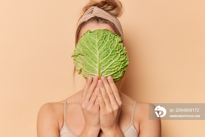 Faceless woman wears headband covers face with cabbage leaf uses natural cosmetics takes care of com