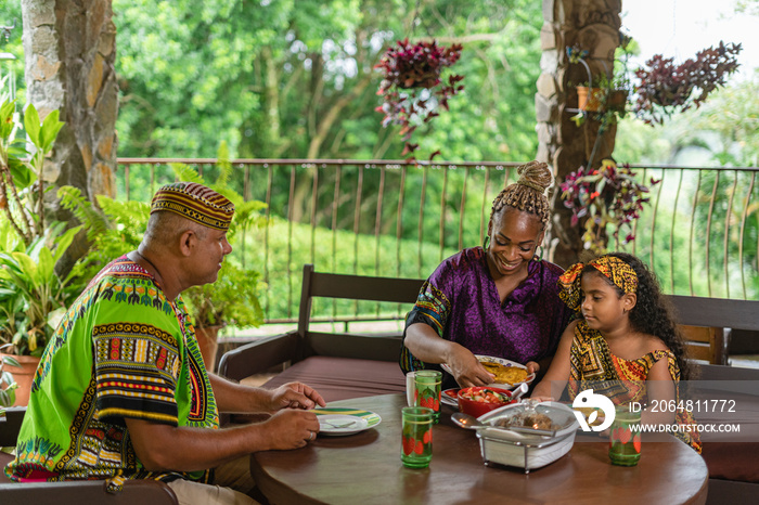 Imagen horizontal de una familia afrocaribeña sentados  juntos a. la mesa sirviéndose un delicioso a