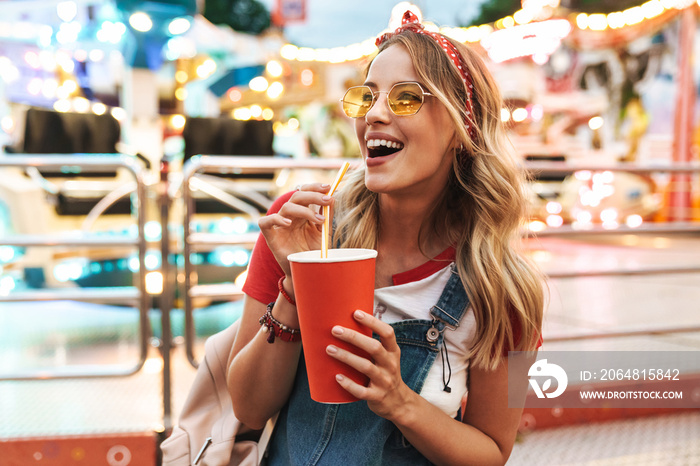 Image of blonde charming woman drinking soda beverage from paper cup while walking in amusement park