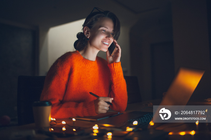 Portrait of happy girl designer working at graphic tablet on laptop talking on smartphone. Wearing e