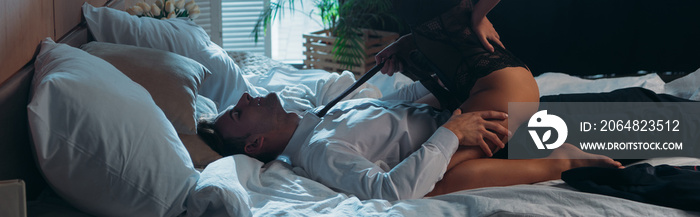 panoramic shot of girlfriend in bodysuit sitting and holding tie of businessman in apartment