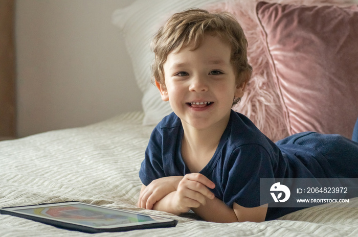 Boy sitting with a tablet in the room. The boy lies on a bed and plays on the tablet.