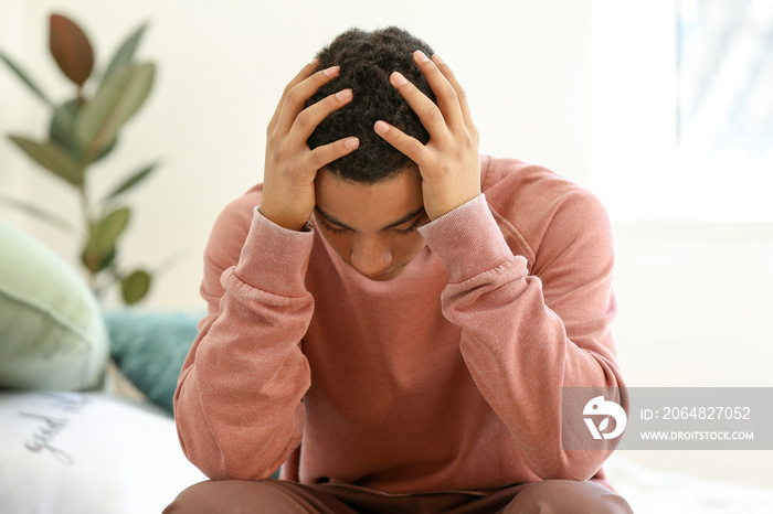 Sad African-American teenage boy sitting on bed at home