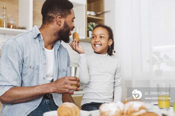 Cheerful black girl feeding her daddy, sitting together at kitchen