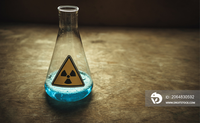 A glass laboratory flask with a blue radioactive liquid on a wooden table. Yellow radiation sign on 