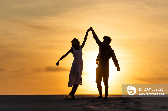 silhouettes of man and woman dancing on beach against sun during sunset