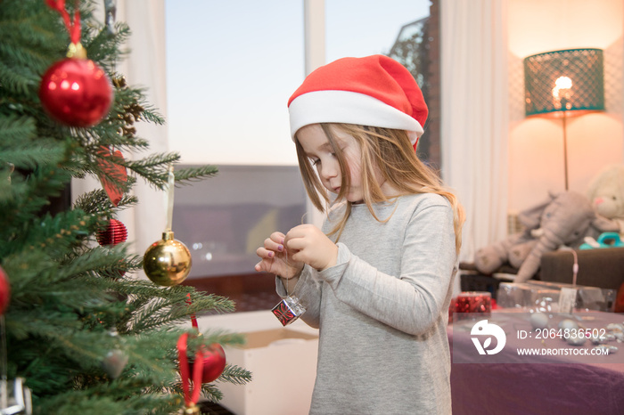 four years blonde cute girl with red Santa Claus hat, decorating Christmas tree at home