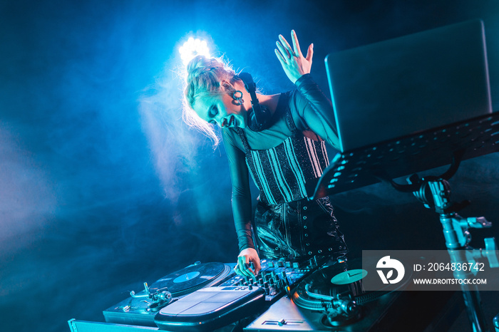 smiling blonde dj girl in headphones standing near dj mixer and gesturing in nightclub with smoke