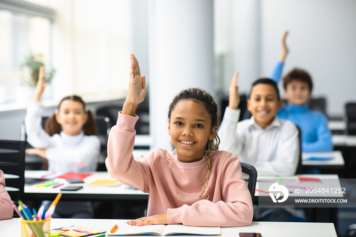 Diverse small schoolkids raising hands at classroom