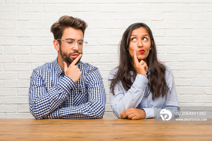 Young indian woman and caucasian man couple doubting and confused, thinking of an idea or worried ab