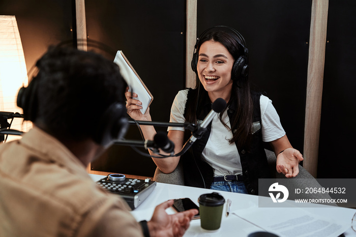 Portait of happy female radio host laughing, listening to male guest, presenter and holding a script