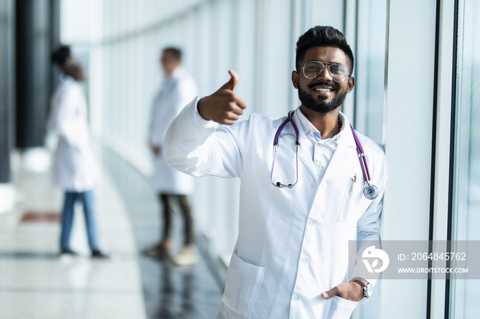 Young indian male doctor with thumbs up in white uniform with collegues on the background