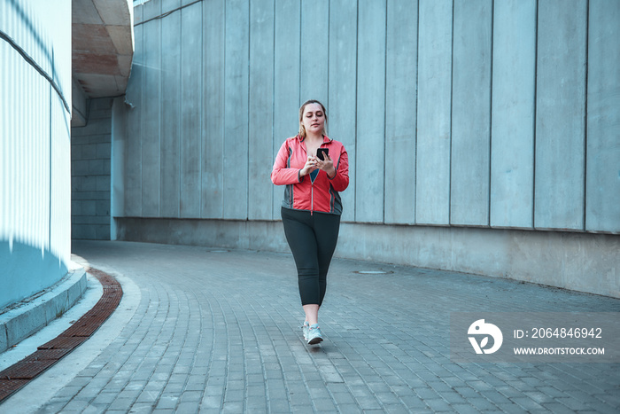 Ready for workout. Young plus size woman on sports clothes holding smart phone while standing outdoo