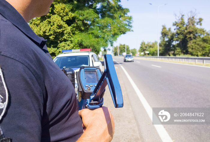 Policeman holding laser speed gun near police car on highway background. Selective focus, part of bo