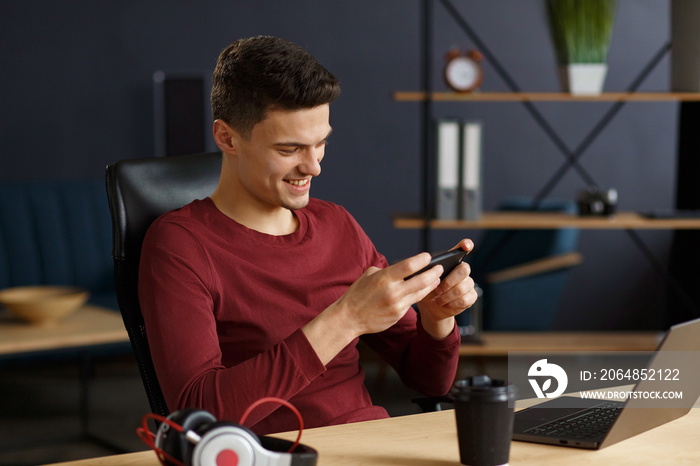 Young man playing games on mobile phone in home office with laptop. Relaxation, taking a break. Youn