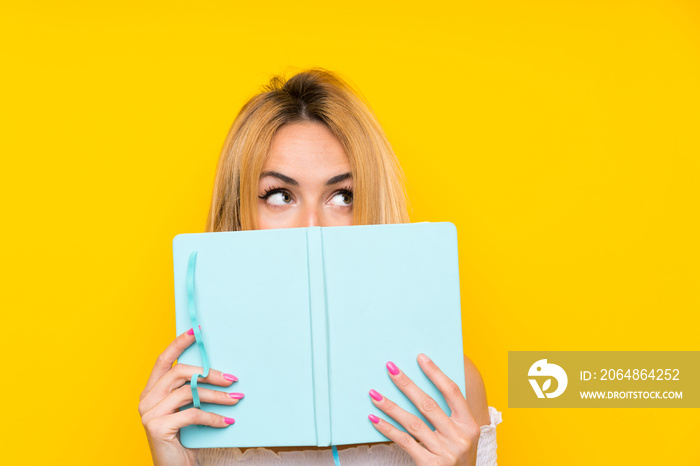 Young blonde woman over isolated yellow wall holding and reading a book