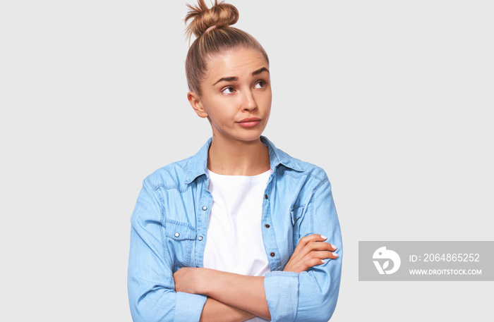 Dissatisfied young woman with crossed hands, wearing casual outfit with hair bun. Unhappy student fe