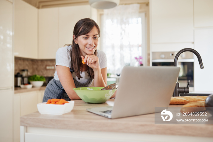 Pretty Caucasian brunette in apron leaning on kitchen counter and looking at laptop and following re