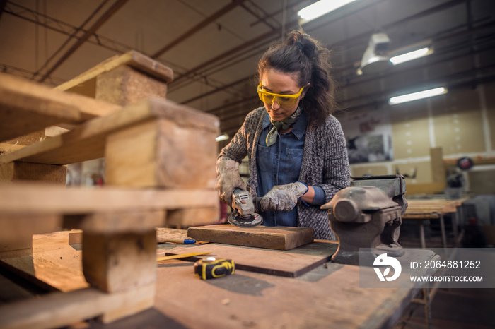 Female carpenter working with sander while standing surrounded with wood.