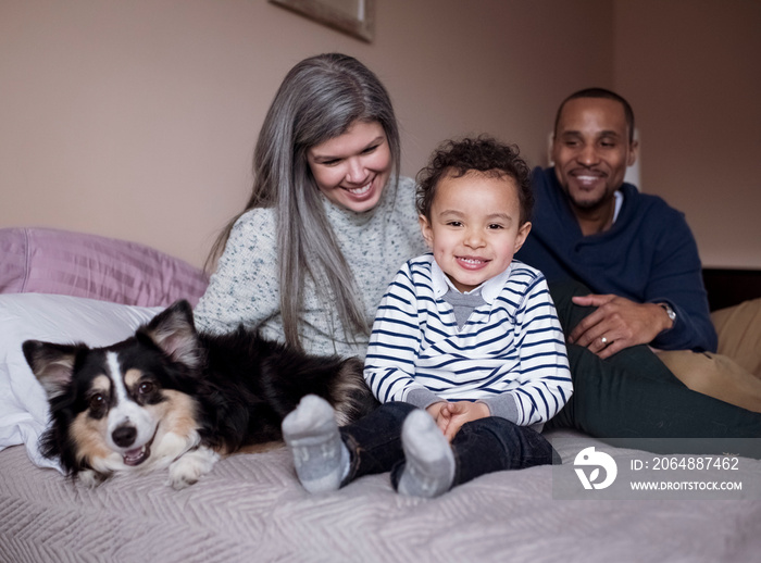 Portrait of cute smiling son sitting with parents and dog on bed against wall at home