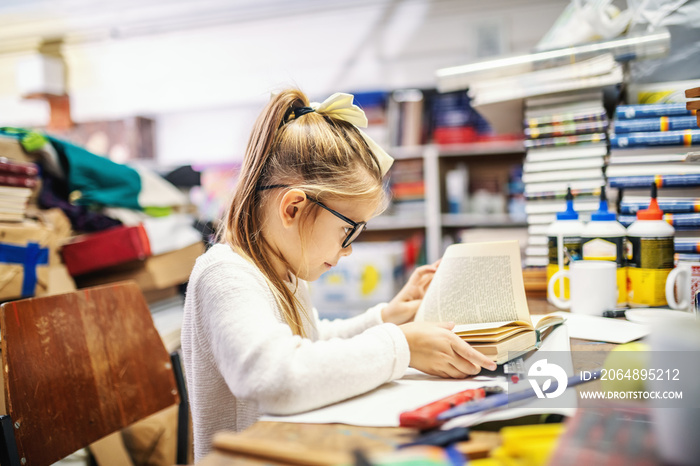 Adorable caucasian blond girl with ponytail and with eyeglasses sitting in bookstore and reading exc