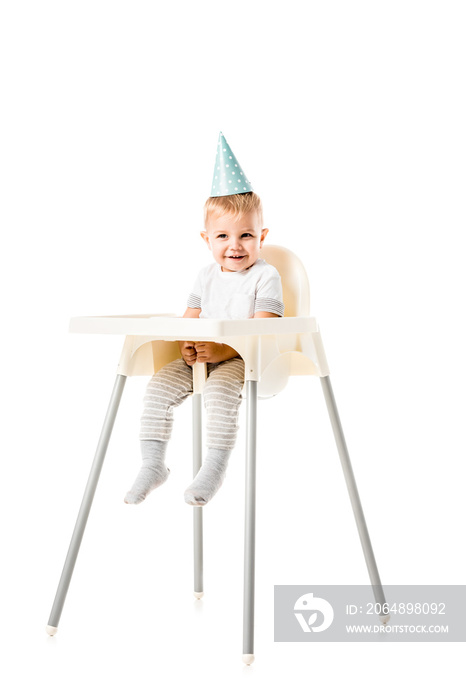 可爱的蹒跚学步的男孩，头戴蓝色派对帽，微笑着坐在白色隔离的高脚椅上