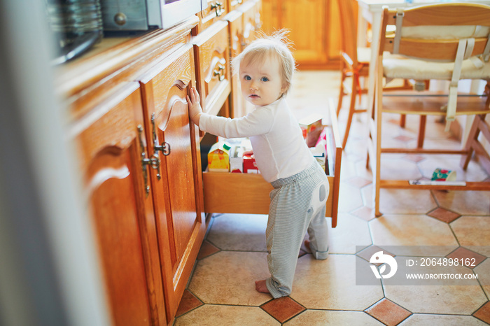 Adorable toddler girl at home, opening the drawer in the kitchen