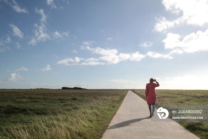 singe woman in red raincoat walking on long straight narrow road amidst green meadows at german nort