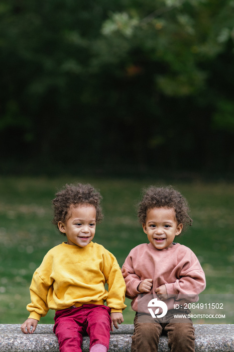 Black and Asian twins sitting on bench in park while smiling