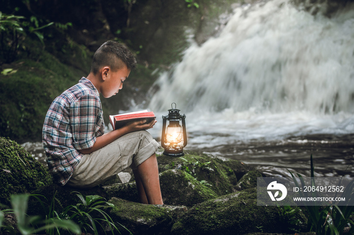 Boy reading book or bible with oil lamp at waterfall. Children and religion.