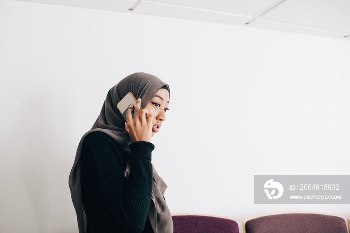 Female student in hijab talking on smart phone while standing in classroom