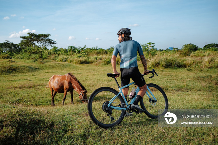 A young bearded cyclist is biking through a field with a horse