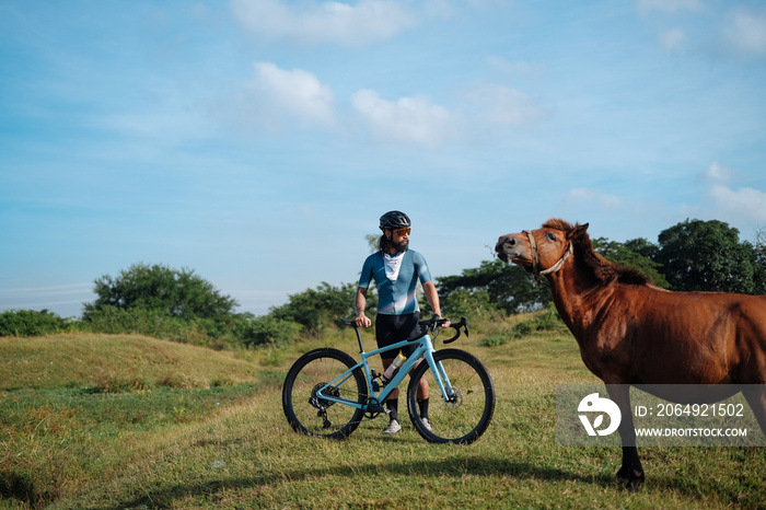 A young bearded cyclist is biking through a field with a horse