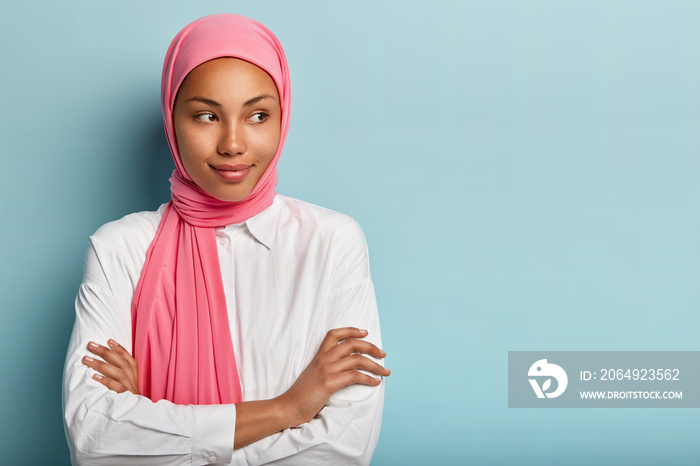 Religious satisfied female model has arms folded, looks aside, has pleased expression, wears pink sc