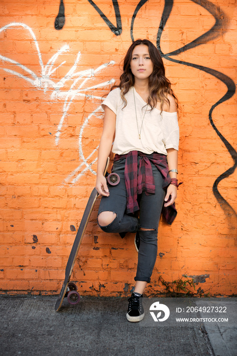 Young female posing with skateboard against brick wall with graffiti