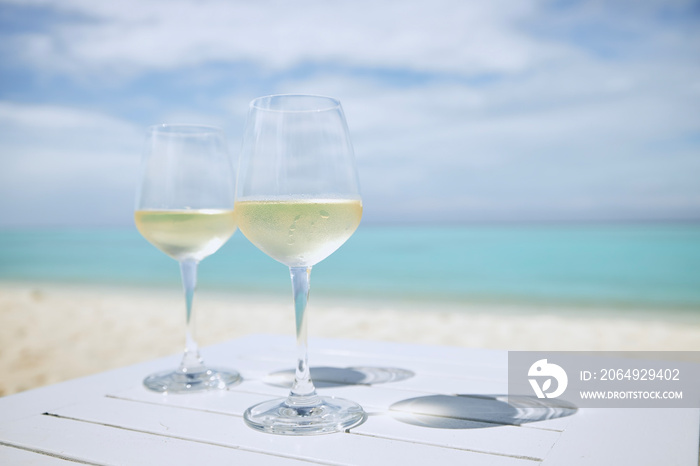 两个装着白葡萄酒的酒杯，放在木桌上，背靠田园诗般的海滩和绿松石般的大海。