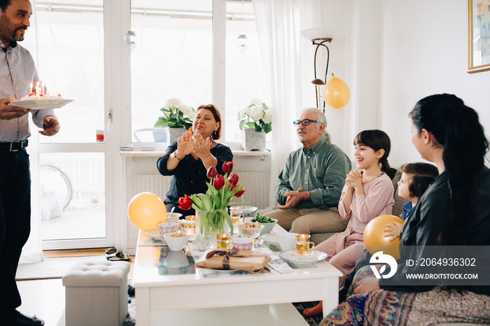 Father with birthday cake walking towards excited family in living room