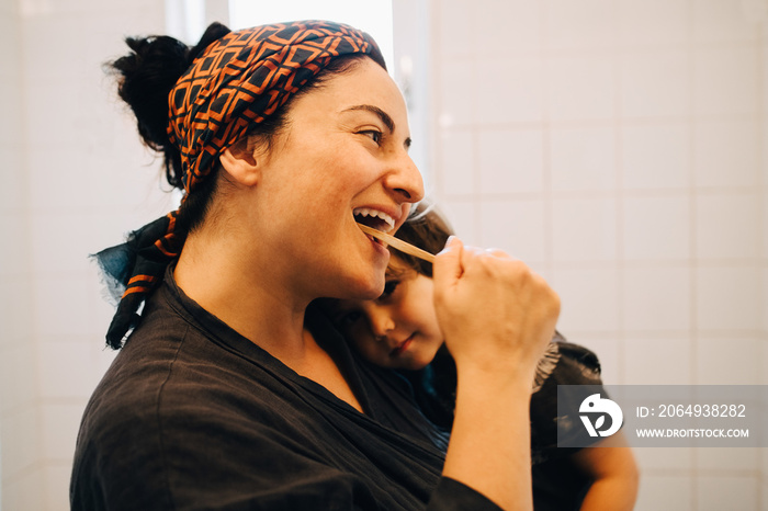 Smiling mother brushing teeth while carrying son at home