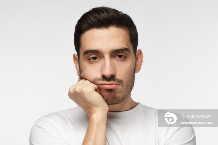 Close up portrait of bored young man in white tshirt with head on chin isolated on grey background