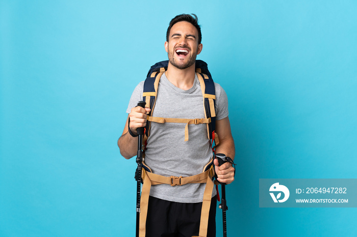 Young caucasian man with backpack and trekking poles isolated on blue background laughing