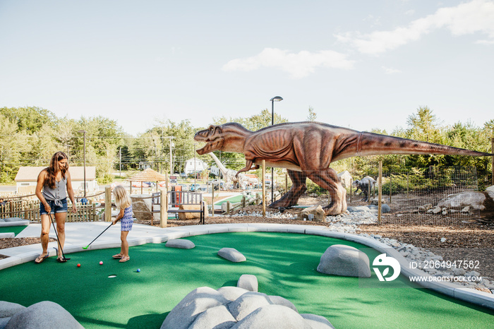 Mother and daughter playing golf against dinosaur sculpture at miniature golf course against sky