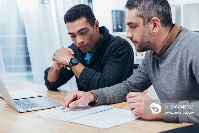 concentrated male coworkers using laptop and working with papers in office