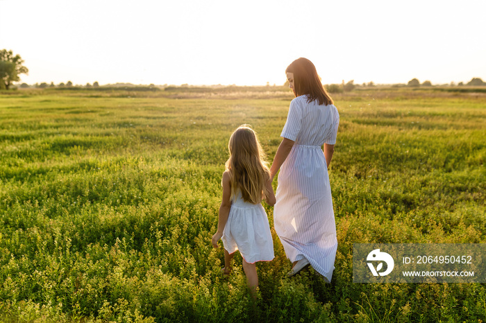 mother and daughter holding hands and walking by green meadow with sunset sky on background