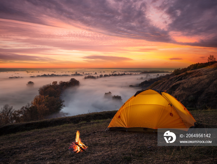 Orange lit inside tent and fire over misty river at sunset