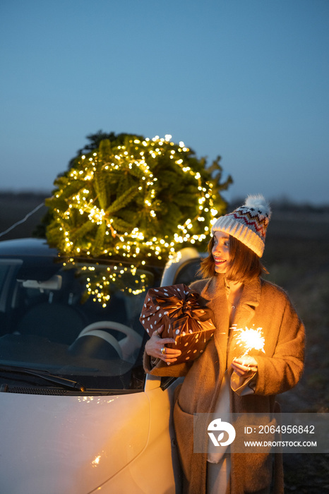 Happy caucasian woman firing sparklers near car with Christmas tree on nature at dusk. Concept of ce