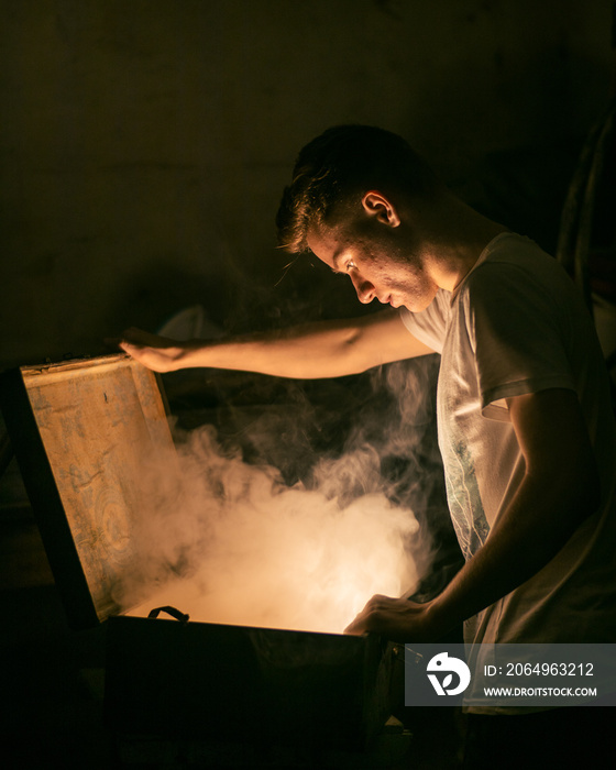 Young man in white t-shirt opening and looking into a chest with smoke and light coming from it